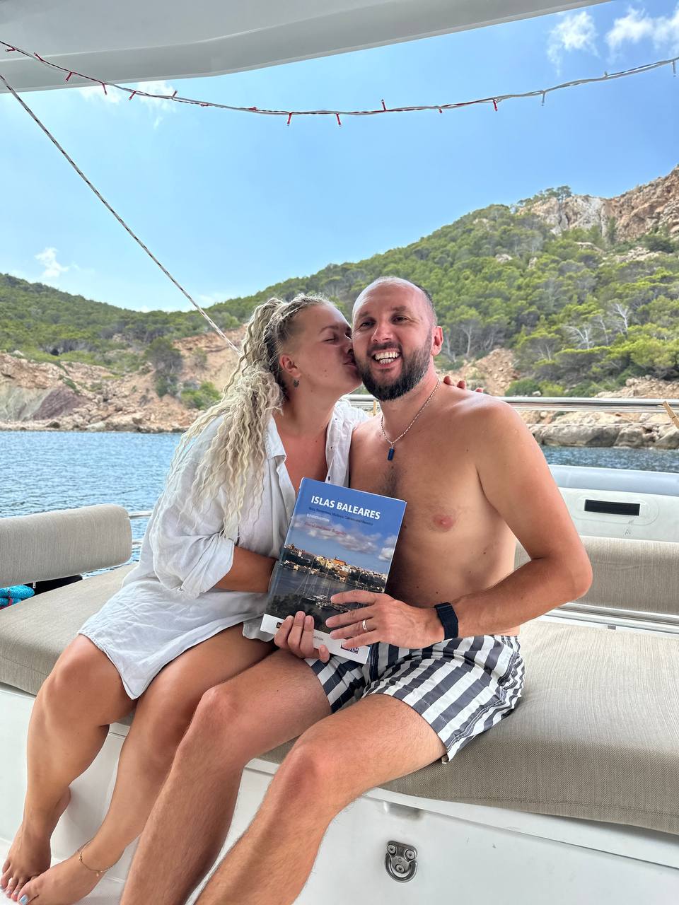 Ana kissing Roman on the cheek while holding a book on the catamaran